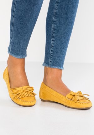 Żółte mokasyny ze skóry zamszowej z frędzlami i kokardką. Podeszwa jest płaska i brązowa. Noszone z niebieskimi dżinsami o postrzępionym obszyciu.