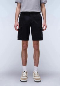 Homem a usar calções cargo pretos, meias brancas, ténis bege e brancos, e uma camisa de manga curta cinza claro, em pé contra um fundo simples.
