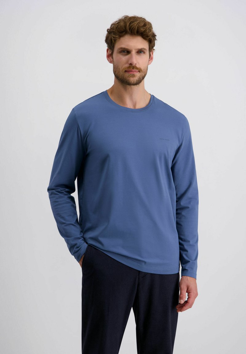 Mann mit lockigem Haar und Bart, der ein blaues langärmliges Hemd und dunkle Hosen trägt, vor einem einfarbigen hellen Hintergrund stehend.