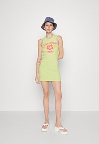Vestido verde em forma de tanque com um design ajustado, apresentando gráficos florais em rosa e o texto "Hanauma Bay". Combinado com um chapéu azul de balde e sandálias brancas de plataforma.