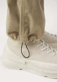 Beżowe spodnie z korduroy z ściąganym mankietem i czarnym elastycznym detalem, połączone z kremowymi sneakersami z zamszu, które mają białe sznurowadła i teksturowaną podeszwę.