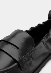 Chaussure à enfiler en cuir noir avec un bout arrondi, présentant une finition lisse, des découpes décoratives et des détails de couture subtils le long des bords.