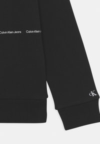 Černá mikina s žebrovanými manžetami a spodním lemem. Nápis "Calvin Klein Jeans" je vytištěn bílou barvou na hrudi. Malé logo "ck" na rukávu.