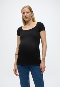 Hosszú szőke hajú, terhes nő, fekete, testhezálló rövid ujjú felsőt és kék farmert visel, egyszínű háttér előtt áll.