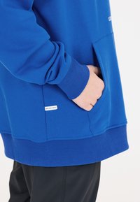 Sweatshirt bleu en tissu doux, avec une poche kangourou, des poignets côtelés et une petite étiquette sur le côté.