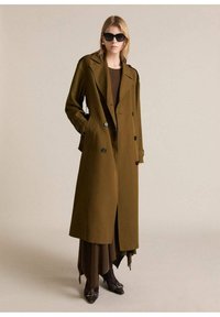 Trench coat de color verde oliva, de doble botonadura, con grandes botones negros, bolsillos laterales y una cintura ceñida con cinturón, superpuesto a un atuendo marrón.