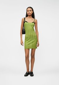 Žena v zelených bezramínkových mini šatech s proužky, černých mokasínech a černé kabelce přes rameno, stojící na čistě bílém pozadí.