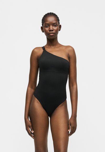 Femme portant un maillot de bain noir asymétrique avec une lanière détaillée, debout devant un fond blanc uni.