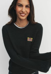 Uśmiechnięta kobieta z długimi ciemnymi włosami nosi czarny sweter z małą kolorową łatą w kształcie kasety na piersi, na jednolitym tle.