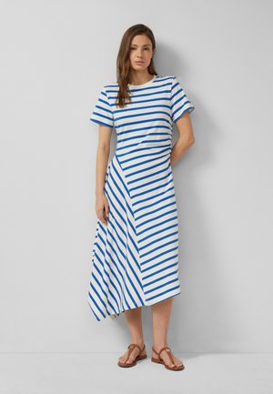 Kobieta w białej sukience midi z niebieskimi poziomymi i ukośnymi paskami, stojąca na tle gładkiej jasnoszarej ściany, na stopach brązowe sandały.