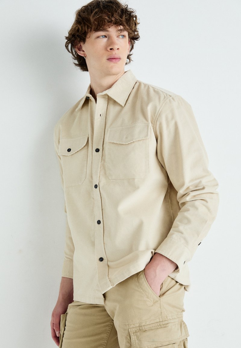 Chemise beige à boutons faite d'un tissu texturé, avec deux poches poitrine, des boutons noirs et des manches longues. Associée à un pantalon cargo beige.