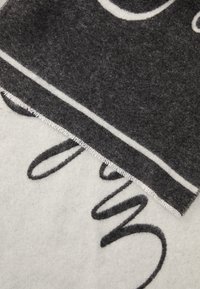 Gros plan sur un tissu doux, frangé, gris et blanc avec un texte en cursive noire partiellement visible sur la section la plus claire.