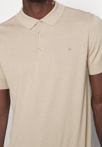 Gros plan d'une personne portant un polo beige à manches courtes avec deux boutons et un petit logo brodé sur la poitrine.