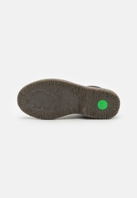 Suela de zapato con superficie de goma texturizada y ranuras, color marrón oscuro, y un distintivo logotipo circular verde incrustado en el diseño.