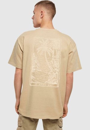 Beige cotton t-shirt featuring a white palm tree graphic on the back and the text "E KOMO MAI" beneath the design. Short sleeves, relaxed fit.