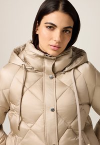 Beige gewatteerde pufferjas met hoge kraag, trekkoordkap en metalen drukknoopsluiting. Heeft een gladde, glanzende textuur.