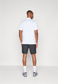 Camisola polo branca de mangas curtas, acompanhada de calções pretos. O modelo está de costas, destacando o design simples e o ajuste da camisola.