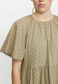 Beige broderet kjole med rund halsudskæring, korte puffærmer og en indsnøret talje med en bølget kant ved ærmerne.