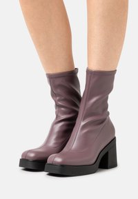 Gambe che indossano stivali in pelle mauve a metà polpaccio con tacco a blocco e suola nera su uno sfondo bianco.