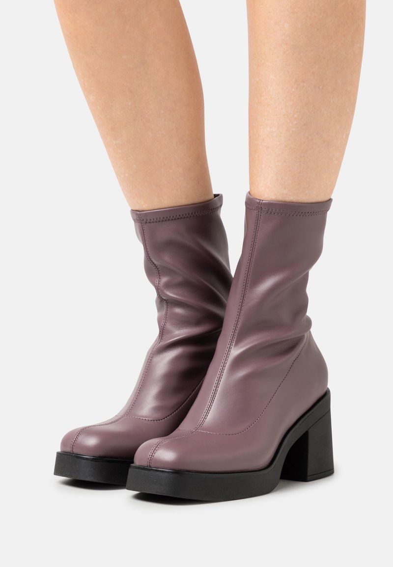 Gambe che indossano stivali in pelle mauve a metà polpaccio con tacco a blocco e suola nera su uno sfondo bianco.