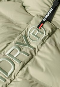 Jasnooliwkowa puchowa kurtka z teksturowanym materiałem, z haftowanym logo "DRY" oraz czarnym/czerwonym uchwytem zamka błyskawicznego z logo.