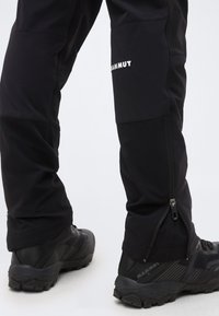 Ben iført sorte Mammut vandrebukser med lynlåse ved anklerne og sorte Mammut trailsko på hvid baggrund.