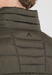 Doudoune en vert olive foncé avec texture matelassée verticale. Elle présente un col haut et un logo noir subtil sur le côté gauche.