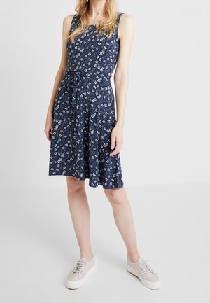 Mujer con vestido azul marino sin mangas hasta la rodilla con pequeño estampado floral blanco y zapatillas blancas, de pie frente a un fondo sencillo.