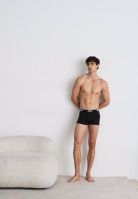 Schwarze Boxershorts mit einem weißen Logo-Bund, getragen von einer barhäuptigen Person. Glatter Stoff und figurbetonte Passform. Sanfter, neutraler Hintergrund.
