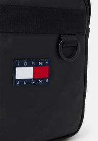 Bolso de hombro de nylon negro con un parche rectangular con franjas rojas, blancas y azules y "TOMMY JEANS" bordado en blanco.