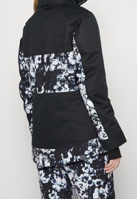 Kvinna iklädd en svart och vit blommönstrad jacka och matchande byxor, stående med ryggen vänd mot kameran.