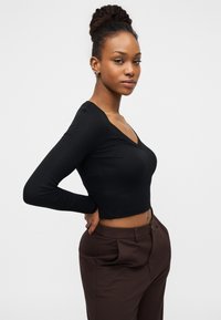 Μαύρο μακρυμάνικο κοντό τοπ με V-λαιμόκοψη, κατασκευασμένο από ελαστικό ύφασμα. Συνδυάζεται με καφέ παντελόνια ψηλού μέσης.