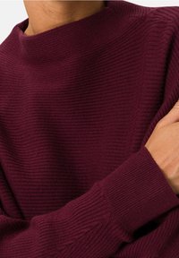 Suéter burdeos con textura acanalada y cuello alto. Presenta mangas largas y ajustadas, además de una tela suave. Primer plano del material.