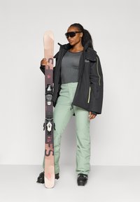 Zwarte waterdichte jas met neonaccenten, aansluitend grijs shirt, mintgroene skibroek, zwarte skibotten en een roze ski met bindingen, rechtop gehouden.