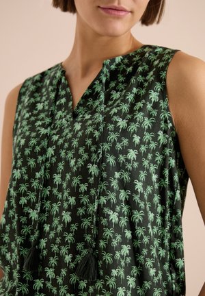 Haut noir sans manches avec motif de palmier vert et liens à pompons à l'avant, porté par une personne aux cheveux bruns courts et au ton neutre sur les lèvres.