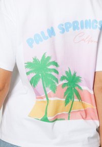 Camiseta blanca con diseño retro en tonos pastel que presenta dos palmeras verdes y el texto "Palm Springs California" en azul y rosa.
