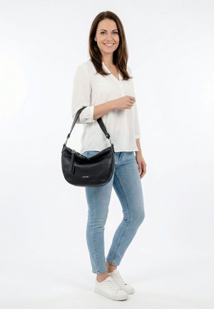 Moteris balta palaidine ir šviesiai mėlynais džinsais stovi laikydama juodos odinės rankinės petį, šypsosi priešais paprastą baltą foną.