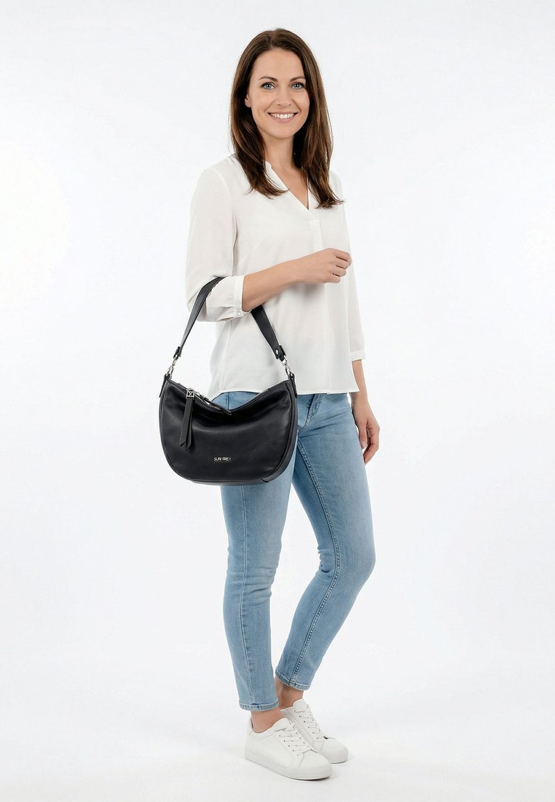 Valge pluusi ja helesiniste teksadega naine seisab, hoides mustast nahast õlakotti, ja naeratab ühevärvilise valge tausta ees.