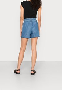 Frau trägt hoch taillierte blaue Jeansshorts, schwarzes ärmelloses Oberteil und schwarze Sandalen, steht mit dem Rücken zur Kamera.