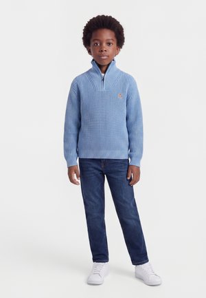 Jeune garçon aux cheveux bouclés foncés, portant un pull bleu à zip, un jean foncé et des baskets blanches, debout sur un fond clair.