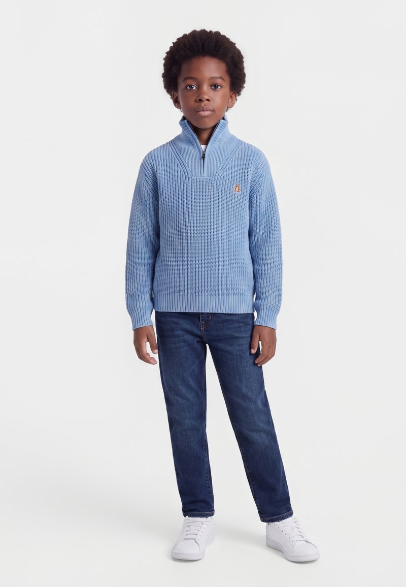 Jeune garçon aux cheveux bouclés foncés, portant un pull bleu à zip, un jean foncé et des baskets blanches, debout sur un fond clair.