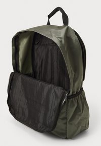 Mochila verde con compartimento principal abierto, forro interior negro con patrones, bolsillos elásticos laterales y asa de transporte acolchada.