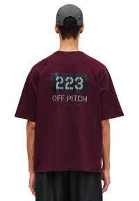 Burgundy oversized t-shirt med en mörk grafisk tryck som har "223" och "OFF PITCH" text på baksidan. Mjukt tyg och avslappnad passform.