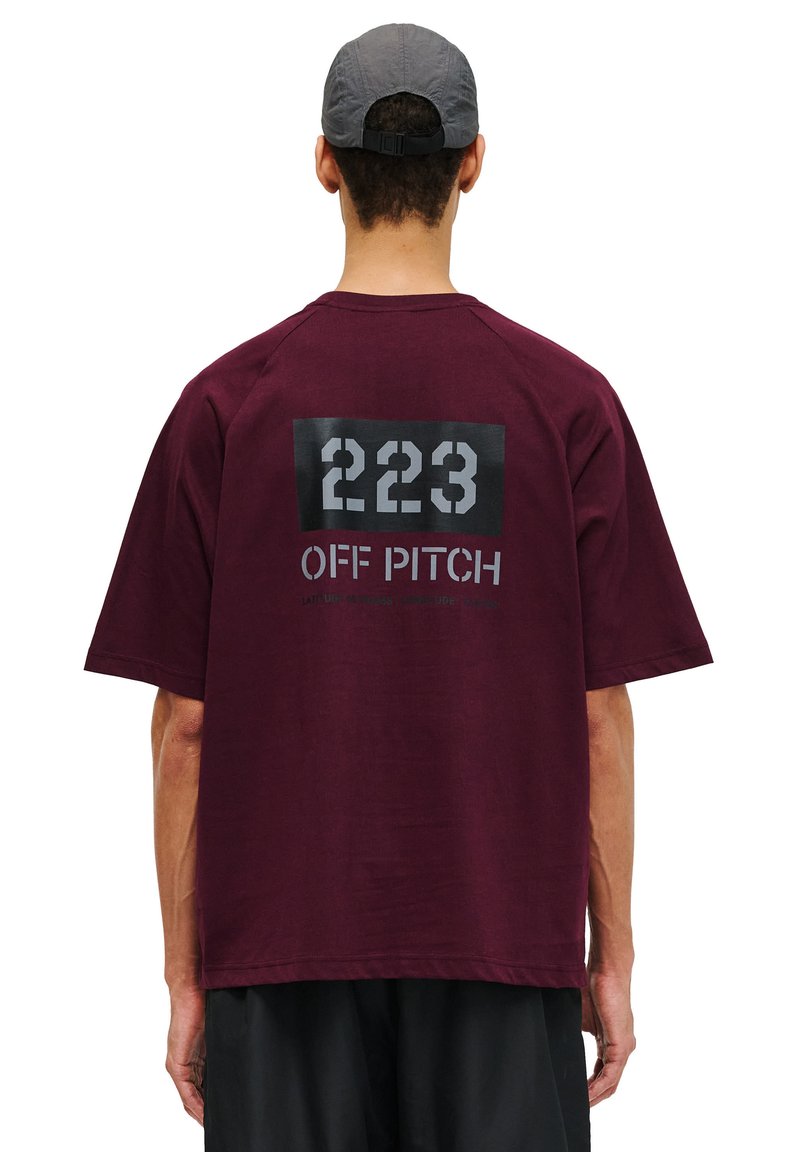 Burgundy oversized t-shirt med en mörk grafisk tryck som har "223" och "OFF PITCH" text på baksidan. Mjukt tyg och avslappnad passform.