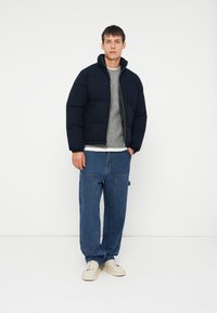 Marine blå pufferjakke med rutete mønster, høy krage og glidelås; båret over en lys grå genser og blå vide jeans.