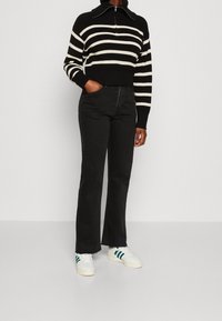 Person trägt schwarze Hochbund-Flared Jeans, einen schwarz-weißen gestreiften Pullover mit Reißverschluss und weiße Sneaker mit grünen Streifen.