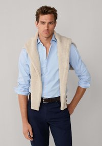 Lichtblauw overhemd met opgerolde mouwen, gecombineerd met een donkere broek. Een crèmekleurige gebreide trui hangt over de schouders.