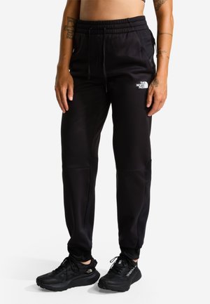 Persoon die zwarte The North Face-broek en zwarte sportschoenen draagt, staand tegen een effen achtergrond.