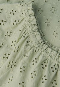 Tessuto di cotone verde chiaro con ricami floreali e fori occhio; presenta una texture arricciata con dettagli a volant lungo il bordo.