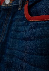 Tummansininen denimmateriaali, jossa on punainen reunus taskun ympärillä, näkyvällä tekstuurilla ja hienovaraisilla kuvioeroilla materiaalissa.